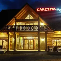Drewniana karczma u Jana o zmierzchu, oświetlona ciepłym światłem. Neonowy szyld z nazwą lśni nad wejściem.