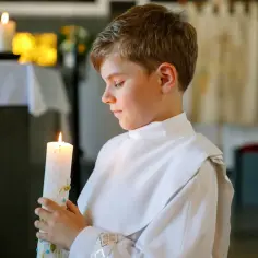 Chłopiec w białej albie trzyma zapaloną świecę, symbolizującą ważny moment, kiedy dzieci idą do komunii.