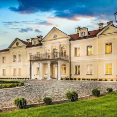Elegancki hotel Sobienie Królewskie w żółtej barwie, z brukowanym podjazdem i zadbanym ogrodem, pod błękitnym niebem.