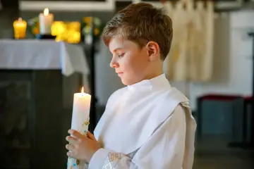 Chłopiec w białej albie trzyma zapaloną świecę, symbolizującą ważny moment, kiedy dzieci idą do komunii.