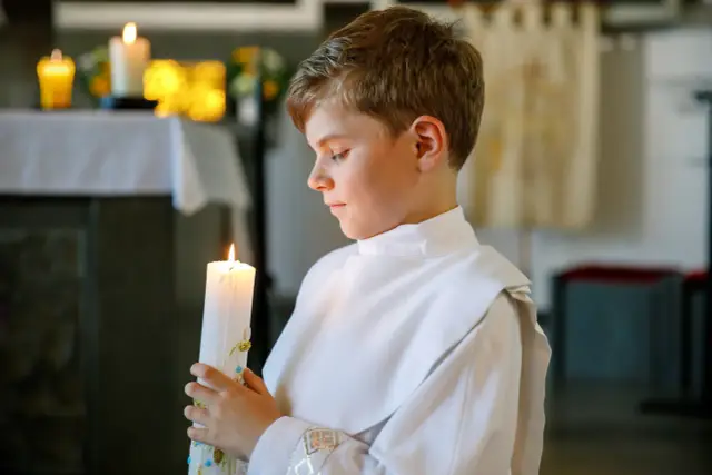 Chłopiec w białej albie trzyma zapaloną świecę, symbolizującą ważny moment, kiedy dzieci idą do komunii.