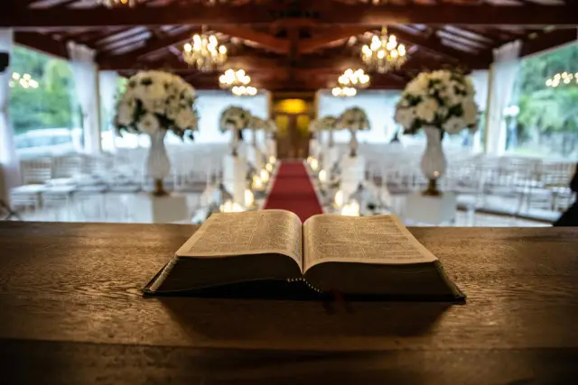 Otwarta Biblia na drewnianym stole. W tle udekorowana sala, gdzie za chwilę rozpocznie się msza. Ile trwa msza w kościele?