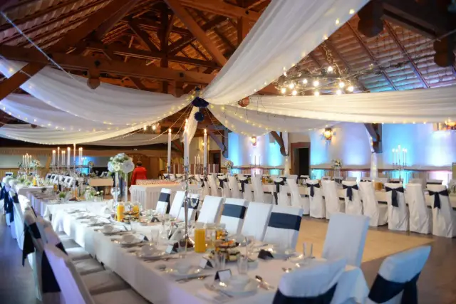 Uroczysta sala weselna w karczmie u Michelle. Długie stoły nakryte białymi obrusami, ozdobione świecami i kwiatami. Krzesła w eleganckich pokrowcach.